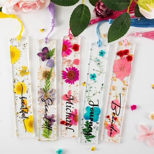 May include: Clear resin bookmarks with dried flowers and gold flakes. Each bookmark has a different flower arrangement and a name written in black script: Beautiful, Scarlett, Michael, Journey, and Brandi. Each has a tassel.