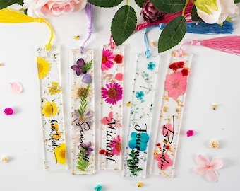Könnte beinhalten: Transparente Harz-Lesezeichen mit getrockneten Blumen und Goldflocken. Jedes Lesezeichen hat ein anderes Blumenarrangement und einen Namen in schwarzer Schrift: Beautiful, Scarlett, Michael, Journey und Brandi. Jedes hat eine Quaste.