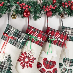 May include: Four white Christmas stockings with plaid cuffs and personalized names in white script. The stockings feature festive appliqués including a green plaid tree, a red plaid snowflake, a white bone, and a white fish skeleton.