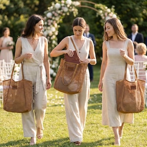 Pode incluir: Tr&ecirc;s pessoas caminhando na grama, cada uma carregando uma grande sacola de malha marrom. Elas est&atilde;o usando macac&otilde;es e vestidos de cores claras. O fundo inclui um arco de casamento, cadeiras e outros convidados, sugerindo um cen&aacute;rio de casamento.