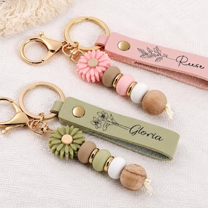 Peut inclure: Deux porte-clés personnalisés avec un anneau et un fermoir en métal doré. L'un possède une lanière en cuir rose avec le nom Reese et un motif de houx, l'autre une lanière vert sauge avec le nom Gloria et un motif floral.
