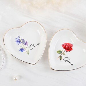 K&ouml;nnte beinhalten: Zwei herzf&ouml;rmige, wei&szlig;e Keramik-Schmuckschalen mit goldenen R&auml;ndern. Eine zeigt eine rote Rose und den Namen "Gloria", die andere blaue und violette Blumen und den Namen "Olive". Eine Perlenkette und eine einzelne Perle befinden sich im Hintergrund.