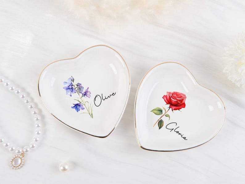 K&ouml;nnte beinhalten: Zwei herzf&ouml;rmige, wei&szlig;e Keramik-Schmuckschalen mit goldenen R&auml;ndern. Eine zeigt eine rote Rose und den Namen "Gloria", die andere blaue und violette Blumen und den Namen "Olive". Eine Perlenkette und eine einzelne Perle befinden sich im Hintergrund.