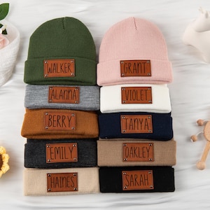 May include: A collection of personalized beanies and headbands in various colors, including green, pink, gray, white, brown, navy, black, and tan. Each item features a leather patch with a name. A sunflower and a toy are also in the image.