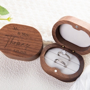 May include: A wooden ring box with a hinged lid, engraved with "Mr. & Mrs. Thomas" and the date "9.15.2025". The box is open, revealing two rings set in a gray velvet interior. The box is oval-shaped.