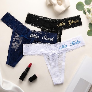 K&ouml;nnte beinhalten: Drei Spitzen-Tangas in Marineblau, Schwarz und Wei&szlig;. Der marineblaue Tanga tr&auml;gt den Schriftzug Mrs Sarah, der schwarze Tanga den Schriftzug Mrs Gloria in Gold und der wei&szlig;e Tanga den Schriftzug Mrs Hester in Blau. Jeder hat eine kleine Schleife.