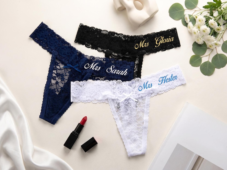 K&ouml;nnte beinhalten: Drei Spitzen-Tangas in Marineblau, Schwarz und Wei&szlig;. Der marineblaue Tanga tr&auml;gt den Schriftzug Mrs Sarah, der schwarze Tanga den Schriftzug Mrs Gloria in Gold und der wei&szlig;e Tanga den Schriftzug Mrs Hester in Blau. Jeder hat eine kleine Schleife.