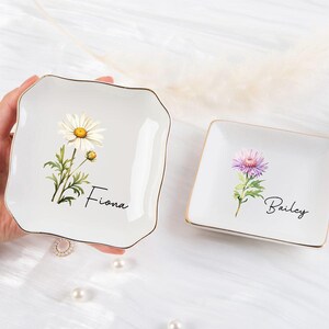 K&ouml;nnte beinhalten: Zwei wei&szlig;e Keramik-Schmuckschalen mit goldenem Rand. Eine quadratische Schale zeigt eine G&auml;nsebl&uuml;mchen-Illustration und den Namen "Fiona". Die andere quadratische Schale hat eine lila Blumenillustration und den Namen "Bailey".
