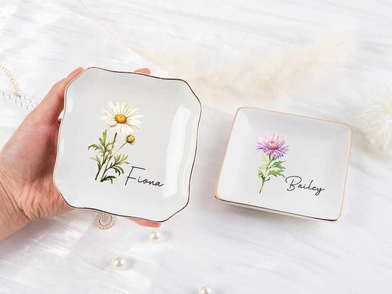 K&ouml;nnte beinhalten: Zwei wei&szlig;e Keramik-Schmuckschalen mit goldenem Rand. Eine quadratische Schale zeigt eine G&auml;nsebl&uuml;mchen-Illustration und den Namen "Fiona". Die andere quadratische Schale hat eine lila Blumenillustration und den Namen "Bailey".