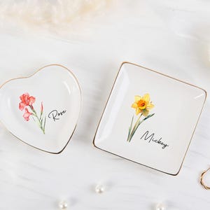 Könnte beinhalten: Zwei weiße Keramik-Schmuckschalen, eine herzförmige und eine quadratische, mit goldenen Rändern. Die Herzschale hat eine rote Blume und den Namen "Rose". Die quadratische Schale hat eine gelbe Narzisse und den Namen "Mickey".