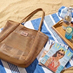 Pode incluir: Uma bolsa tote de malha bege com um patch de couro que diz "Emilia" est&aacute; sobre uma toalha de praia listrada azul e branca. Uma bandeja de madeira cont&eacute;m uma garrafa de &aacute;gua, um copo de l&iacute;quido azul com uma fatia de lim&atilde;o, uma ma&ccedil;&atilde;, um lim&atilde;o e fatias de abacate. Uma revista tamb&eacute;m est&aacute; presente.