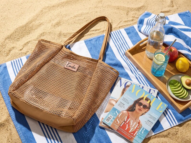 Pode incluir: Uma bolsa tote de malha bege com um patch de couro que diz "Emilia" est&aacute; sobre uma toalha de praia listrada azul e branca. Uma bandeja de madeira cont&eacute;m uma garrafa de &aacute;gua, um copo de l&iacute;quido azul com uma fatia de lim&atilde;o, uma ma&ccedil;&atilde;, um lim&atilde;o e fatias de abacate. Uma revista tamb&eacute;m est&aacute; presente.
