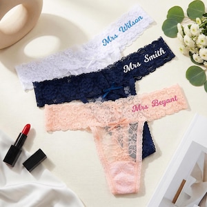 K&ouml;nnte beinhalten: Drei Spitzen-Tangas in Wei&szlig;, Marineblau und Pfirsich, jeweils mit einer kleinen Schleife in der Mitte. Der wei&szlig;e Tanga ist mit Mrs Wilson, der marineblaue mit Mrs Smith und der pfirsichfarbene mit Mrs Beyant bestickt.