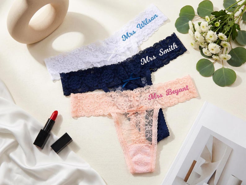 K&ouml;nnte beinhalten: Drei Spitzen-Tangas in Wei&szlig;, Marineblau und Pfirsich, jeweils mit einer kleinen Schleife in der Mitte. Der wei&szlig;e Tanga ist mit Mrs Wilson, der marineblaue mit Mrs Smith und der pfirsichfarbene mit Mrs Beyant bestickt.