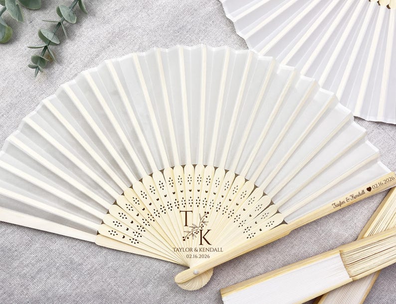 May include: White folding fan with a wooden handle. The fan features the initials "T K" with floral accents and the names "Taylor & Kendall" and the date "02.16.2026" engraved on the handle. The fan is partially open, displaying its pleated design.