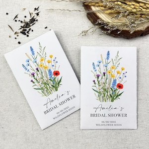 Op de afbeelding: Twee witte enveloppen met wilde bloemenzaad. Elke envelop heeft een bloemenontwerp met blauwe, gele, rode en groene bloemen. De tekst op de enveloppen luidt "Amelia's Bridal Shower 06/08/2025 Wildflower Seeds".