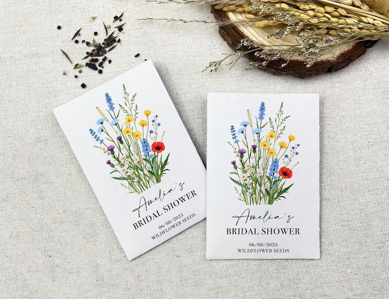 Op de afbeelding: Twee witte enveloppen met wilde bloemenzaad. Elke envelop heeft een bloemenontwerp met blauwe, gele, rode en groene bloemen. De tekst op de enveloppen luidt "Amelia's Bridal Shower 06/08/2025 Wildflower Seeds".