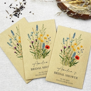 Op de afbeelding: Drie kraftpapieren enveloppen met zakjes met wilde bloemenzaad. Elke envelop heeft een bloemenontwerp en de tekst "Amelia's Bridal Shower" met de datum "06/08/2023" en de woorden "Wildflower Seeds".