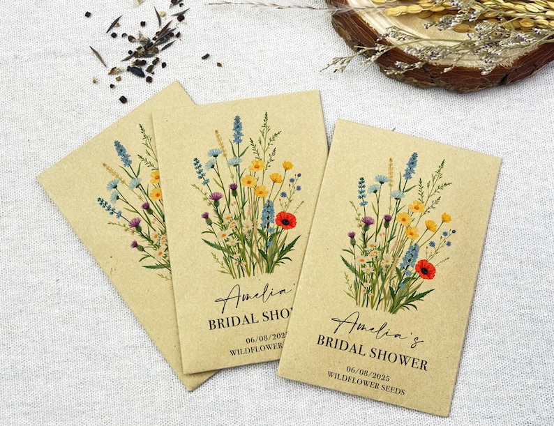 Op de afbeelding: Drie kraftpapieren enveloppen met zakjes met wilde bloemenzaad. Elke envelop heeft een bloemenontwerp en de tekst "Amelia's Bridal Shower" met de datum "06/08/2023" en de woorden "Wildflower Seeds".