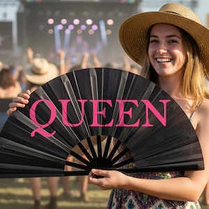 Może przedstawiać: Czarny wachlarz z różowym napisem "QUEEN". Wachlarz trzymany jest przez osobę w słomkowym kapeluszu, uśmiechającą się przed tłumem na imprezie plenerowej.