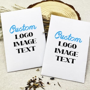 Könnte beinhalten: Zwei weiße Karten mit dem Wort "Custom" in hellblauer Schrift oben, gefolgt von "LOGO IMAGE TEXT" in schwarzen Blockbuchstaben. Die Karten liegen auf einer neutralfarbenen Oberfläche mit dekorativen Elementen.