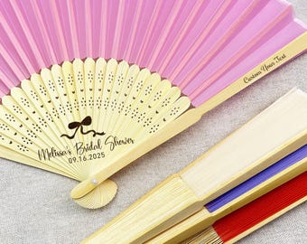 May include: A collection of decorative hand fans in various colors. One fan is open, displaying a pink and cream design with the text "Melissa's Bridal Shower 09.16.2025" and a bow graphic. Other fans are closed, showing red, blue, and cream colors.