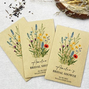 Op de afbeelding: Drie kraftpapieren enveloppen met zakjes met wilde bloemenzaad. Elke envelop heeft een bloemenontwerp en de tekst "Amelia's Bridal Shower" met de datum "06/08/2023" en de woorden "Wildflower Seeds".
