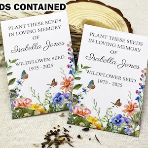 May include: Two white seed packets with floral designs. The text on the packets reads "Plant these seeds in loving memory of Isabella Jones Wildflower Seed 1975-2025".