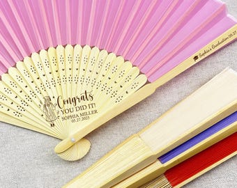 May include: Pink and cream graduation fan with the text "Congrats YOU DID IT!" and the graduate's name and date. Additional fans with red, blue, and cream accents are also visible.