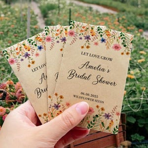 May include: Seed packets with floral designs and the text "Let Love Grow" and "Amelia's Bridal Shower" are held in a hand. The packets are tan with colorful flowers and the date 06.08.2025.