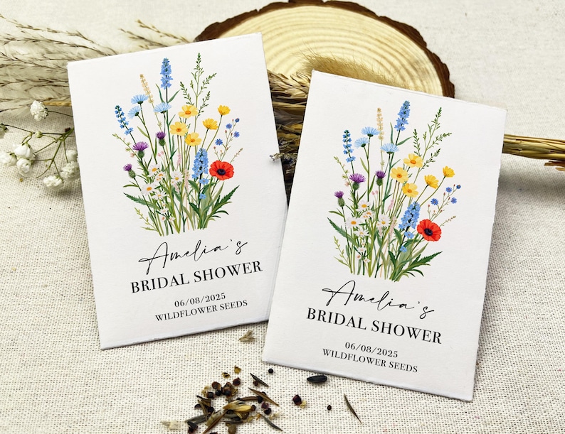 Op de afbeelding: Twee witte zaadzakjes met een bloemenmotief en de tekst "Amelia's Bridal Shower" en "06/08/2025 Wildflower Seeds".
