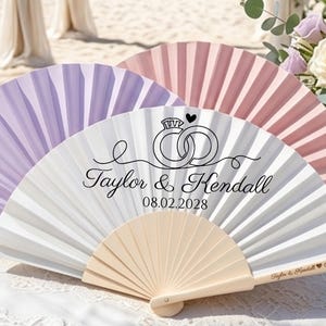 Puede incluir: Tres abanicos plegables en blanco, lavanda y rosa claro. El abanico blanco presenta un gráfico negro de dos anillos entrelazados con un corazón, el texto Taylor & Kendall y la fecha 08.02.2028. El mango es de madera clara con texto.