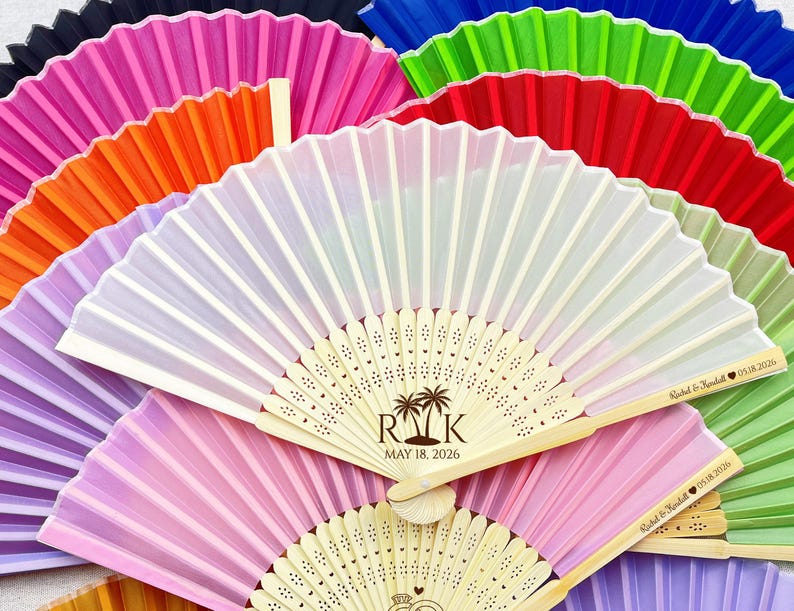May include: A collection of colorful, folded paper fans in various shades, including pink, orange, green, and blue. Some fans have personalized text and dates, with wooden handles and decorative cutouts. Ideal for weddings or events.
