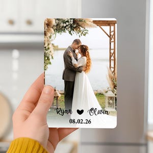Puede incluir: Impresión fotográfica rectangular con una pareja abrazándose bajo un arco floral. La impresión incluye los nombres "Ryan" y "Olivia", un símbolo de corazón y la fecha "08.02.26". La imagen es sostenida por una persona.