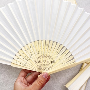 May include: White folding fan with a bamboo handle and a floral design. The fan is personalized with the names "Sophie & Wyatt" and the date "05.16.2026". The fan is partially open, displaying its pleated design.