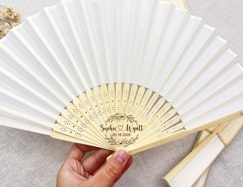 May include: White folding fan with a bamboo handle and a floral design. The fan is personalized with the names "Sophie & Wyatt" and the date "05.16.2026". The fan is partially open, displaying its pleated design.