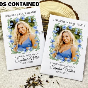 May include: Two white seed packets with blue floral designs and a photo of a person. The text on the packets reads "Forever in our hearts" and "In loving memory of Sophia Miller 1995-2025".