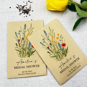 Op de afbeelding: Twee kraftpapieren enveloppen met zakjes wilde bloemenzaad. De enveloppen hebben een bloemenontwerp met verschillende kleuren, waaronder blauw, geel, rood en groen. De tekst op de enveloppen luidt "Amelia's Bridal Shower" en "06/08/2025 Wildflower Seeds".