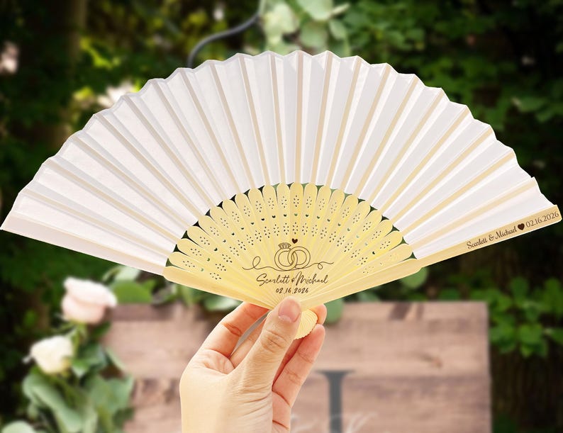 May include: A white folding fan with a light wood handle. The fan features a decorative design with wedding rings and the names "Scarlett & Michael" and the date "02.16.2026". The fan is held open, displaying its pleated design.