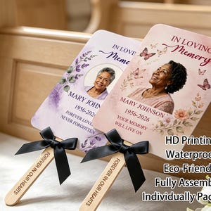 May include: Two rectangular memorial hand fans with wooden handles and black ribbon bows. Each fan features a portrait of Mary Johnson, the text IN LOVING MEMORY, 1956-2026, and floral graphics. The handles are engraved with FOREVER IN OUR HEARTS.