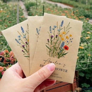 Op de afbeelding: Drie bruine papieren enveloppen met wilde bloemenzaadzakjes. De enveloppen hebben een bloemenontwerp met blauwe, gele en rode bloemen. De tekst op de envelop luidt "Amelia's Bridal Shower Flower Seeds".