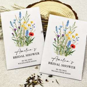 Op de afbeelding: Twee witte zaadzakjes met een bloemenmotief en de tekst "Amelia's Bridal Shower" en "06/08/2025 Wildflower Seeds".