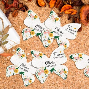 May include: Butterfly-shaped memorial cards with floral designs and the name "Olivia" and dates 1945-2025. The cards also feature the text "Forever In Our Hearts". The cards are on a corkboard surface.