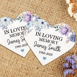 May include: Two heart-shaped memorial plaques with floral designs in shades of purple and blue. The plaques read "IN LOVING MEMORY" and "James Smith 1985-2025". A small blue heart is at the bottom of each plaque. The background is a textured, tan surface.