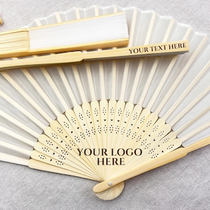 May include: A collection of folding fans with bamboo frames and white fabric. One fan is open, displaying a decorative design and the text "YOUR LOGO HERE." Another fan is closed, with the text "YOUR TEXT HERE." These fans are ideal for weddings or special events.
