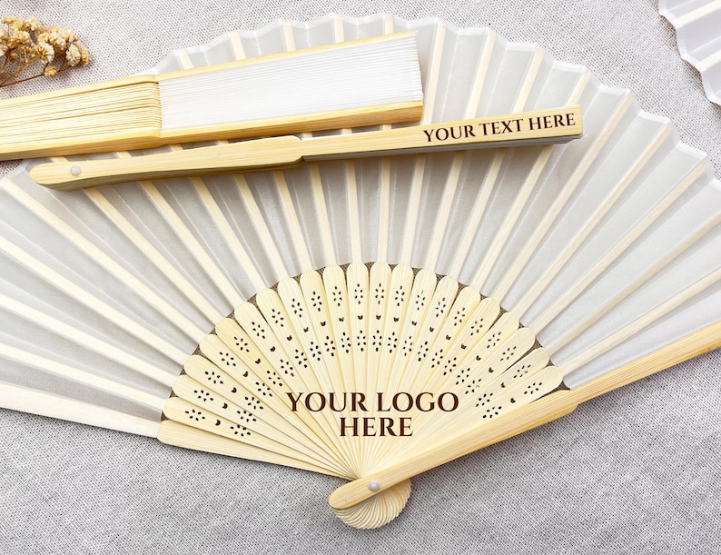 May include: A collection of folding fans with bamboo frames and white fabric. One fan is open, displaying a decorative design and the text "YOUR LOGO HERE." Another fan is closed, with the text "YOUR TEXT HERE." These fans are ideal for weddings or special events.