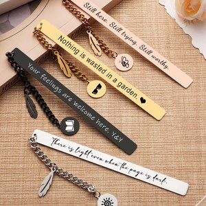 May include: A collection of metal bookmarks in various colors, including black, gold, and rose gold. Each bookmark features an inspirational quote and a decorative charm, such as a feather or a sun. The bookmarks are displayed on a wooden surface.