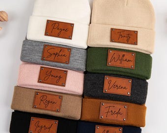 May include: A collection of ten knit beanies in various colors including white, beige, gray, pink, tan, black, olive green, dark gray, brown, and navy blue. Each beanie features a rectangular brown leather patch with a custom name engraved.