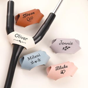May include: A black stethoscope with a white personalised leather ID tag labelled Oliver, surrounded by four additional leather tags in tan, grey, blue, and pink, each engraved with names Steven, Jennie, Milani, and Blake alongside small icons.