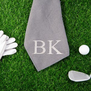 May include: A gray golf towel with the initials "BK" embroidered in white. The towel is lying on green grass next to a white golf ball, a golf club, and a pair of white golf gloves.